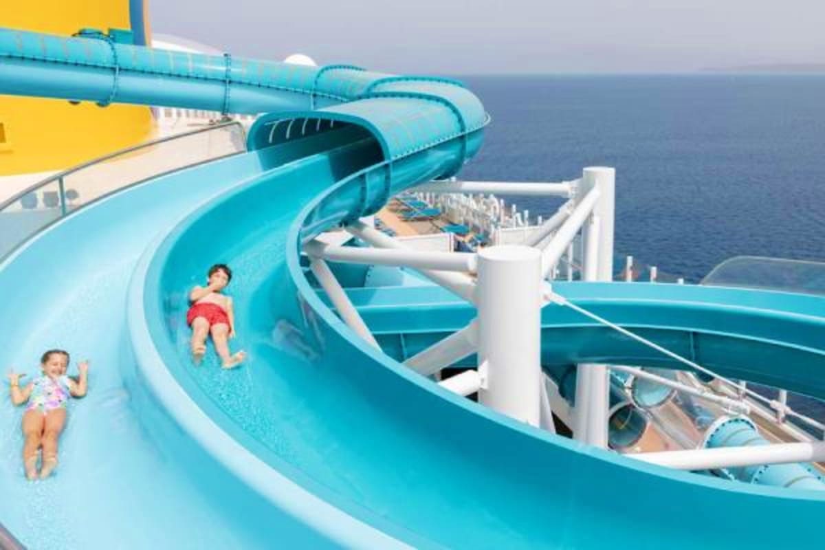 Children sliding down bright turquoise water slides on a cruise ship deck with ocean visible in background